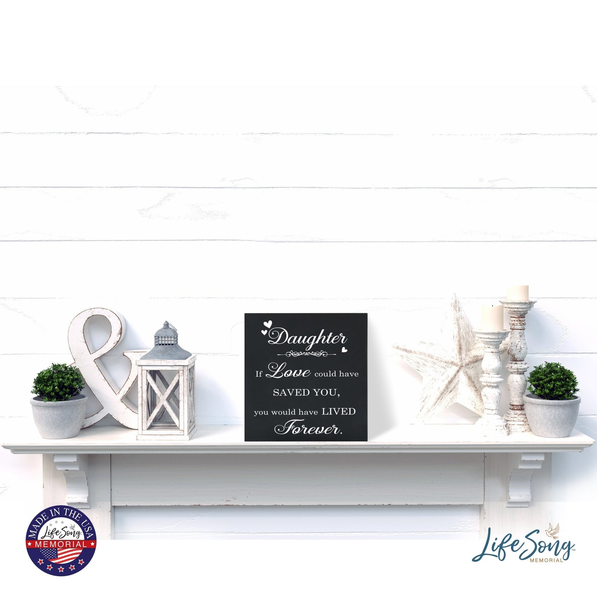 A decorative white shelf showcases a LifeSong Milestones Memorial Shadow Box Urn for human ashes, adorned with delightful accents such as lanterns, greenery, and star-shaped decorations. The urn features heartfelt engraving, fostering a serene and comforting ambiance.